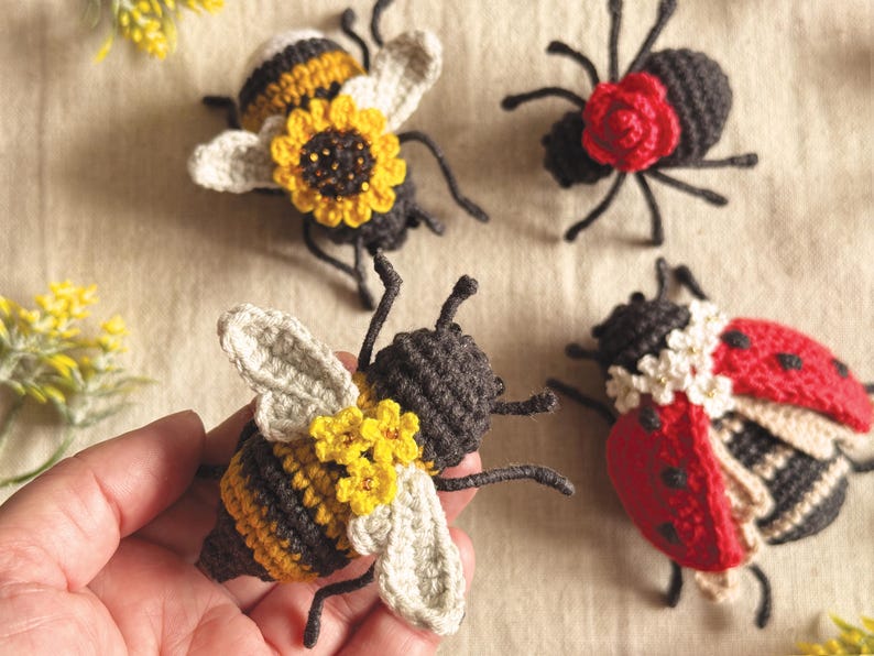 May include: Crocheted insect ornaments, including a bee with yellow and black stripes, a ladybug with red and black wings, and a spider with a red flower. The bee has yellow flower accents and the ladybug has white accents.