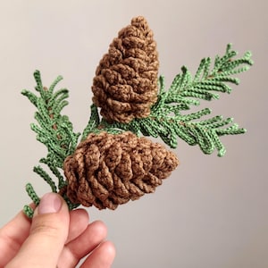May include: A crochet decoration featuring two brown pinecones attached to a green coniferous branch. The pinecones have a textured, layered appearance, and the branch has detailed, needle-like foliage crafted with fine green yarn.