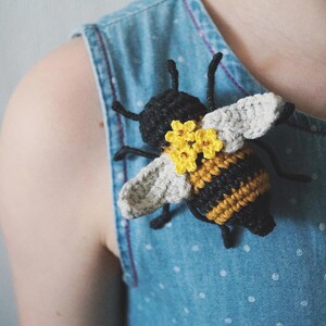 May include: A crocheted bee brooch featuring a black and yellow striped body, light gray wings, and three small yellow flowers attached to the back. The bee has thin black antennae and legs, and is pinned to a light blue denim fabric with white dots.