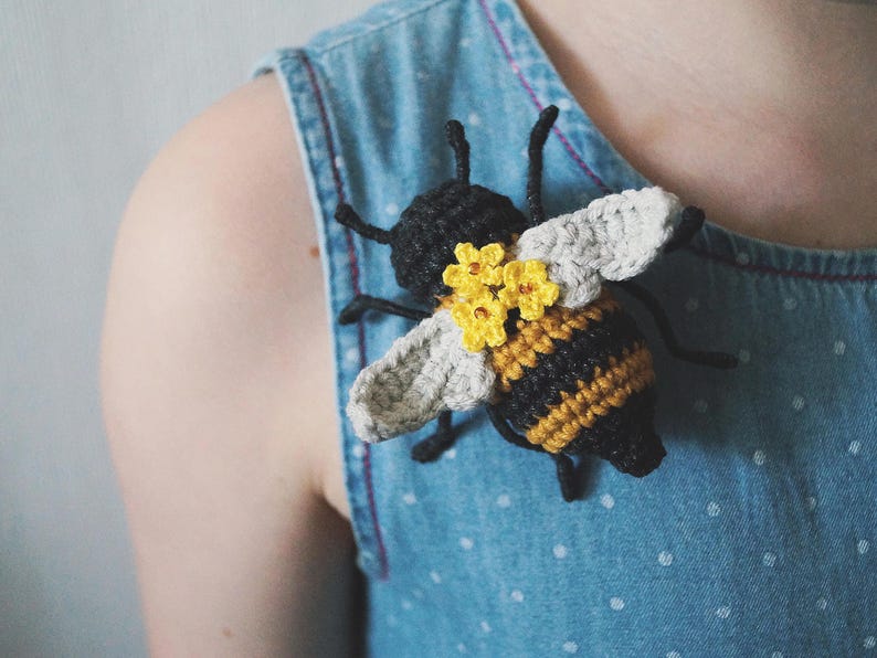 May include: A crocheted bee brooch featuring a black and yellow striped body, light gray wings, and three small yellow flowers attached to the back. The bee has thin black antennae and legs, and is pinned to a light blue denim fabric with white dots.
