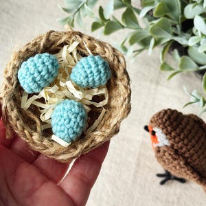 May include: A handmade crochet nest with three light blue eggs and a small brown crochet bird with white and orange accents. The nest is made of woven twine and filled with straw.