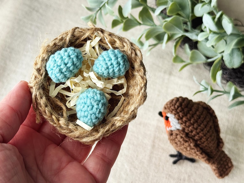 May include: A handmade crochet nest with three light blue eggs and a small brown crochet bird with white and orange accents. The nest is made of woven twine and filled with straw.