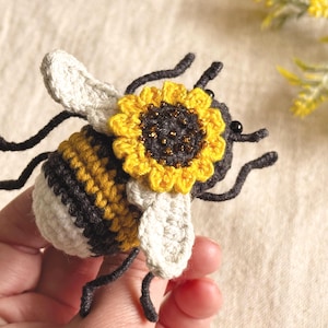Puede incluir: Una abeja de ganchillo con un girasol en la espalda. La abeja tiene una cabeza blanca, un cuerpo a rayas amarillas y negras, y patas y antenas negras. El girasol es amarillo con un centro oscuro y está sujeto a la espalda de la abeja.