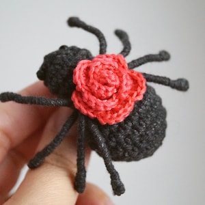 May include: A small, black crocheted spider featuring a textured body and eight thin legs. A vibrant, coral-red crocheted rose is attached to the top of the spider's back, providing a distinct contrast to the dark yarn used for the rest of the figure.