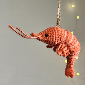 May include: A handmade, orange crochet shrimp ornament with white stripes. The shrimp has black eyes and long, thin antennae. It is suspended by a natural twine string, perfect for hanging. The background is a soft, neutral color with a few out-of-focus lights.