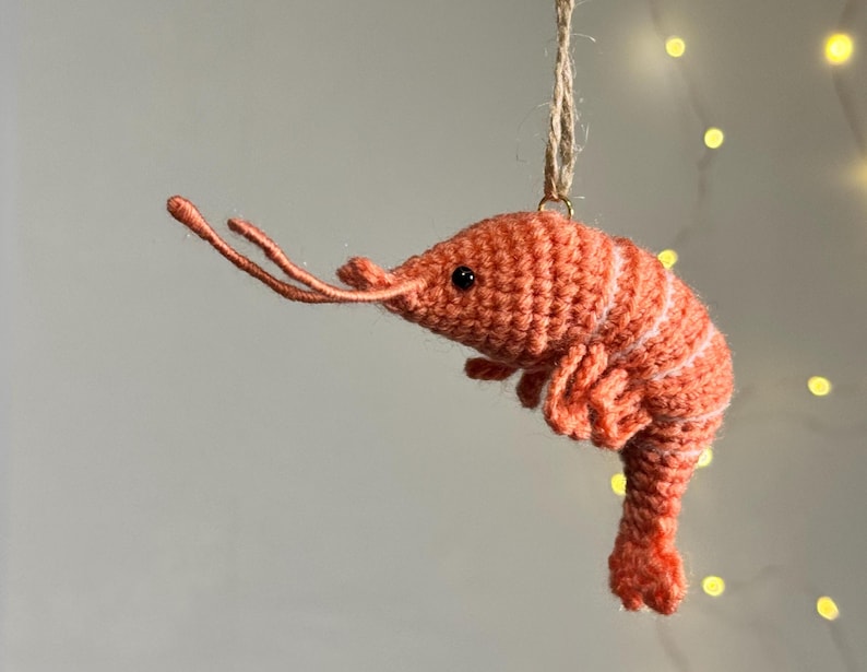 May include: A handmade, orange crochet shrimp ornament with white stripes. The shrimp has black eyes and long, thin antennae. It is suspended by a natural twine string, perfect for hanging. The background is a soft, neutral color with a few out-of-focus lights.