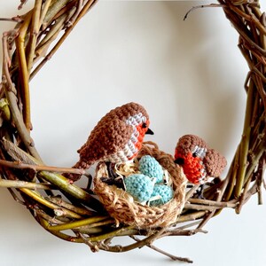 May include: A handcrafted wreath made of woven twigs, featuring two crocheted robins and a nest with three blue eggs. The birds have brown and white bodies with red breasts. The wreath is a charming decorative piece.