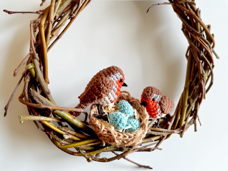 May include: A handcrafted wreath made of woven twigs, featuring two crocheted robins and a nest with three blue eggs. The birds have brown and white bodies with red breasts. The wreath is a charming decorative piece.