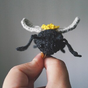 May include: A small, handcrafted crochet bee figurine featuring a textured black body, two light gray wings, and a bright yellow flower detail on its back. The insect has two black bead eyes and thin, dark, articulated legs extending outward.