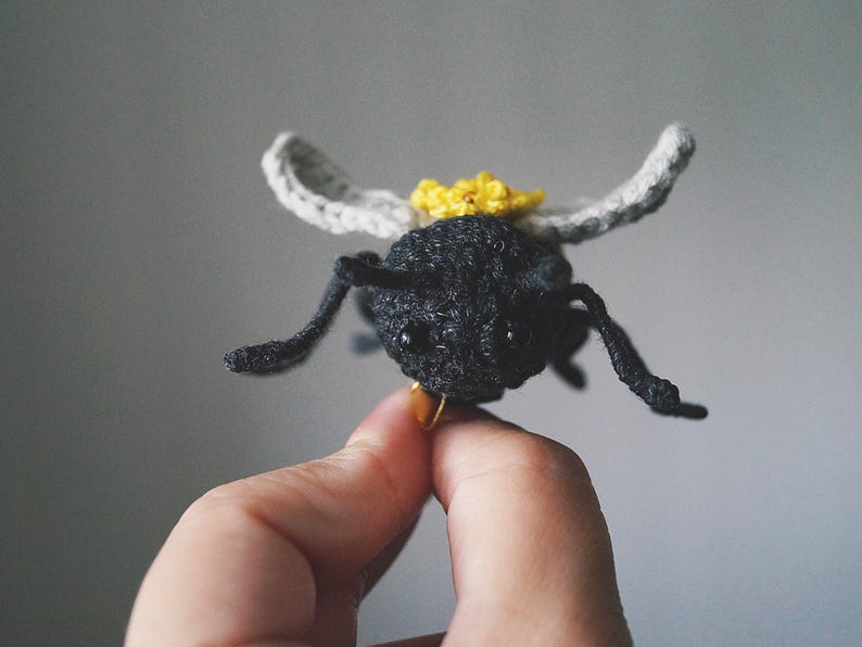 May include: A small, handcrafted crochet bee figurine featuring a textured black body, two light gray wings, and a bright yellow flower detail on its back. The insect has two black bead eyes and thin, dark, articulated legs extending outward.