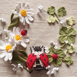 Könnte beinhalten: Ein Häkelarrangement mit weißen Gänseblümchen mit gelber Mitte, grünen vierblättrigen Kleeblättern und kleinen weißen Blüten. Ein großer roter Marienkäfer mit schwarzen Punkten und schwarzem Körper sitzt unten, ein kleinerer auf einem Gänseblümchen.