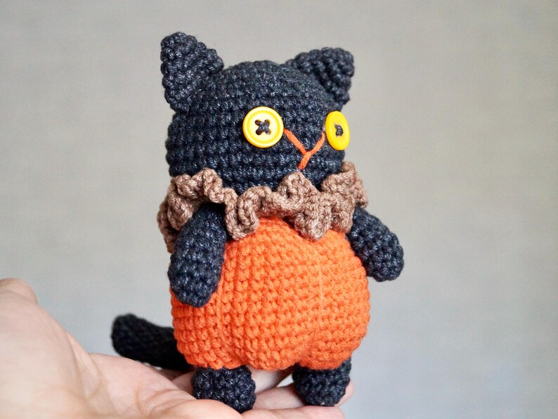 May include: A handmade crochet cat doll with a black head and limbs, orange body, and brown ruffled collar. The cat has yellow button eyes and a small black tail. A unique, whimsical plush toy.
