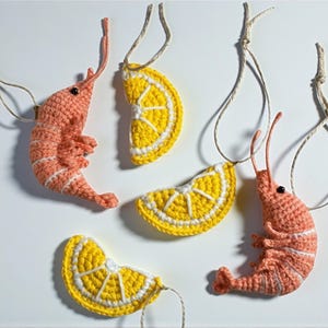 May include: Crocheted ornaments featuring a shrimp and lemon slice design. The shrimp are coral with white stripes, and the lemon slices are yellow with white details. Each ornament has a string for hanging.