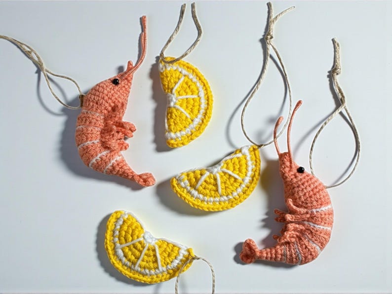 May include: Crocheted ornaments featuring a shrimp and lemon slice design. The shrimp are coral with white stripes, and the lemon slices are yellow with white details. Each ornament has a string for hanging.
