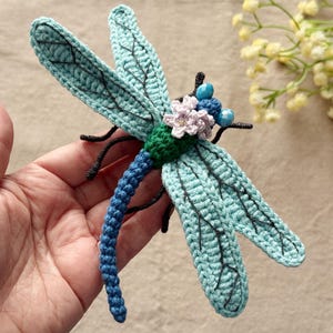 Peut inclure: Une libellule au crochet faite à la main, dans des tons de bleu, vert et noir. La libellule présente des ailes détaillées, un corps texturé et des ornements floraux. Cet objet fantaisiste est un article artisanal unique.