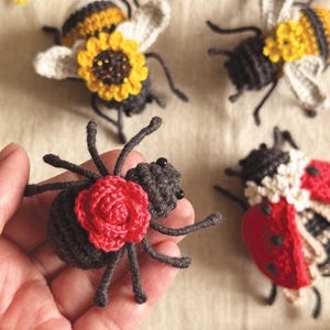 May include: A collection of four handmade crochet insects including two bees with yellow sunflower accents, one ladybug with white flower details, and one dark grey spider featuring a prominent red crocheted rose on its back.