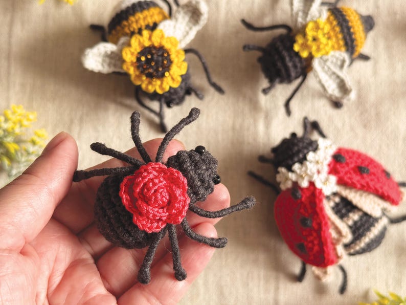 May include: A collection of four handmade crochet insects including two bees with yellow sunflower accents, one ladybug with white flower details, and one dark grey spider featuring a prominent red crocheted rose on its back.
