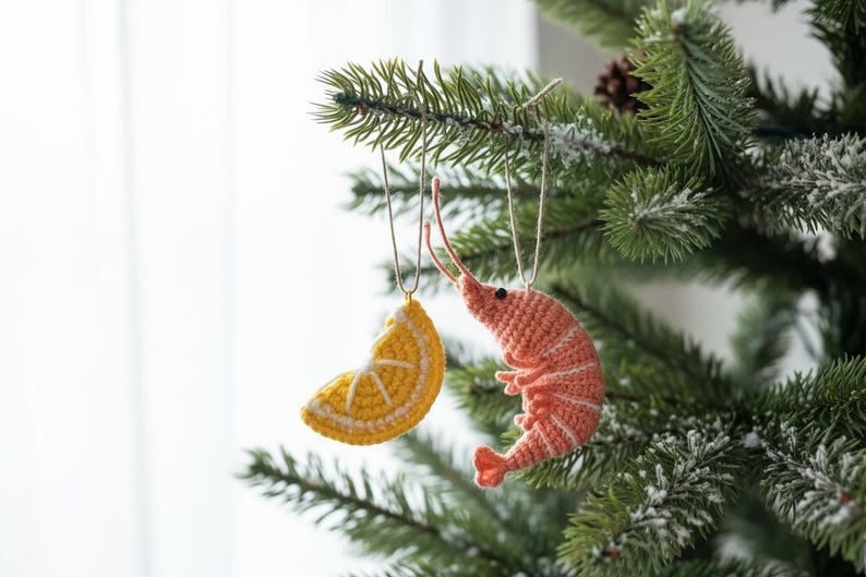 May include: Two handmade Christmas ornaments hanging on a tree. One is a yellow crocheted lemon slice, and the other is a pink crocheted shrimp. The ornaments are suspended by silver wire and white string.