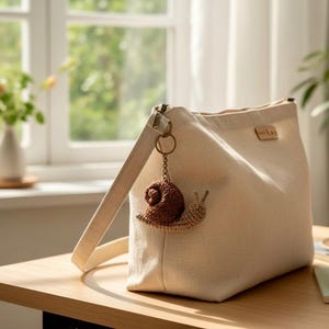 May include: A cream-colored canvas shoulder bag featuring a brown crocheted snail keychain attached to the side strap. The bag has a rectangular silhouette with a small rectangular brand label near the top edge and a sturdy shoulder strap.