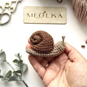 May include: A small, handmade crochet snail featuring a dark brown coiled shell and a light beige body with two small antennae. The snail is crafted with visible yarn stitches, showcasing a textured, soft appearance in a compact, handheld size.