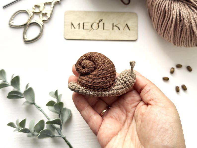 May include: A small, handmade crochet snail featuring a dark brown coiled shell and a light beige body with two small antennae. The snail is crafted with visible yarn stitches, showcasing a textured, soft appearance in a compact, handheld size.