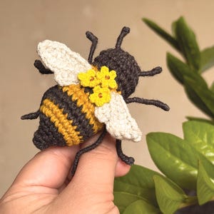 May include: A crocheted bee toy featuring a dark brown and yellow striped body, off-white wings, and three small yellow flowers attached to the back. The bee has six dark brown legs and two antennae, crafted with detailed yarn stitching throughout.