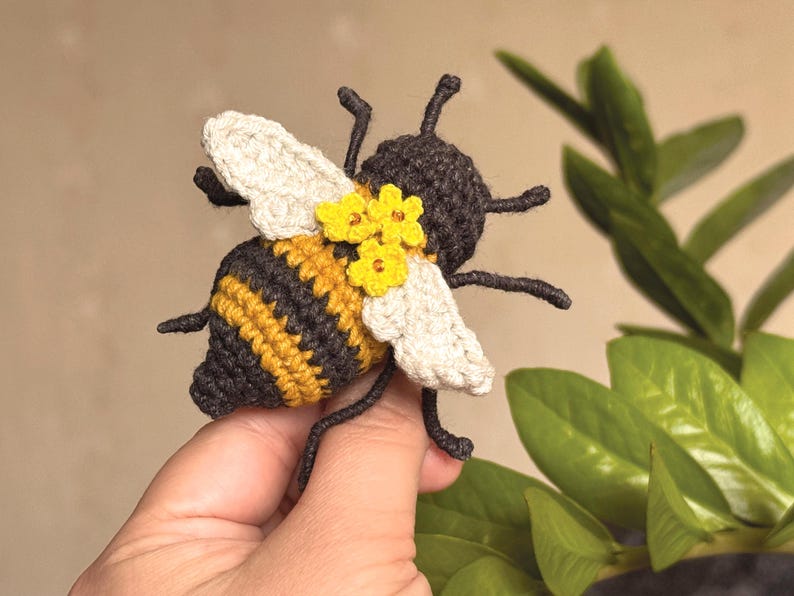 May include: A crocheted bee toy featuring a dark brown and yellow striped body, off-white wings, and three small yellow flowers attached to the back. The bee has six dark brown legs and two antennae, crafted with detailed yarn stitching throughout.