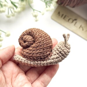 May include: A small, handmade crochet snail featuring a dark brown, spiraled shell and a light beige body with two upright antennae. The snail is crafted with visible yarn stitches, showcasing a textured, soft appearance and a compact design.