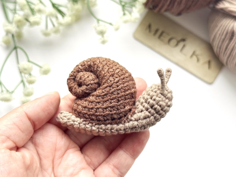 May include: A small, handmade crochet snail featuring a dark brown, spiraled shell and a light beige body with two upright antennae. The snail is crafted with visible yarn stitches, showcasing a textured, soft appearance and a compact design.