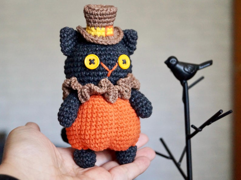 May include: A handmade crochet cat doll with a black body, orange pumpkin-shaped belly, and a brown top hat. The cat has yellow button eyes and a brown ruffled collar. A black metal bird decoration is in the background.