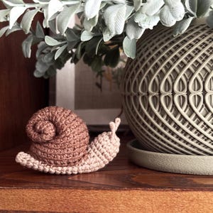 May include: A small, handmade crochet snail figurine featuring a light beige body and a textured, spiraled brown shell. The snail has two small antennae on its head and is crafted with visible yarn stitches, showcasing a soft, tactile appearance.