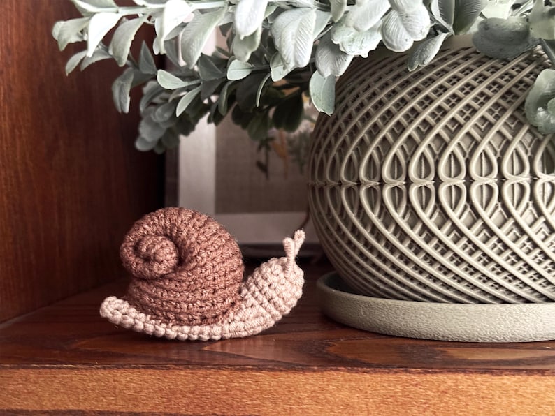 May include: A small, handmade crochet snail figurine featuring a light beige body and a textured, spiraled brown shell. The snail has two small antennae on its head and is crafted with visible yarn stitches, showcasing a soft, tactile appearance.