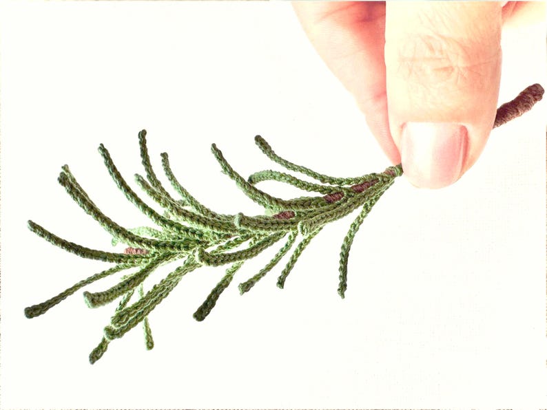 Op de afbeelding: Een handgemaakte gehaakte tak in groen- en bruintinten, die lijkt op een botanisch element. De tak wordt door een hand tegen een witte achtergrond gehouden. Het haakwerk is gedetailleerd en gestructureerd.