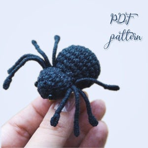 Op de afbeelding: Een gehaakte zwarte spin met acht poten en kleine zwarte ogen. De spin is handgemaakt en lijkt een speelgoed of decoratief item te zijn. De tekst "PDF pattern" is zichtbaar.