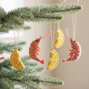 May include: Crocheted Christmas tree ornaments in the shape of shrimp and lemon slices. The shrimp are coral-colored, and the lemon slices are yellow with white details. The ornaments hang from a green Christmas tree.