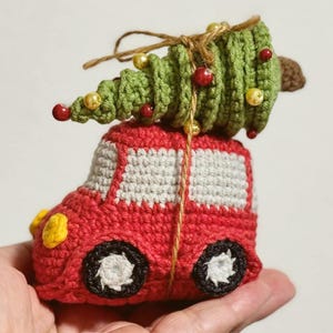 Può includere: Un ornamento natalizio fatto a mano all'uncinetto. Presenta un'auto rossa con dettagli di finestre bianche e ruote nere. Un albero di Natale verde all'uncinetto con ornamenti di perline rosse e dorate è legato all'auto con dello spago.