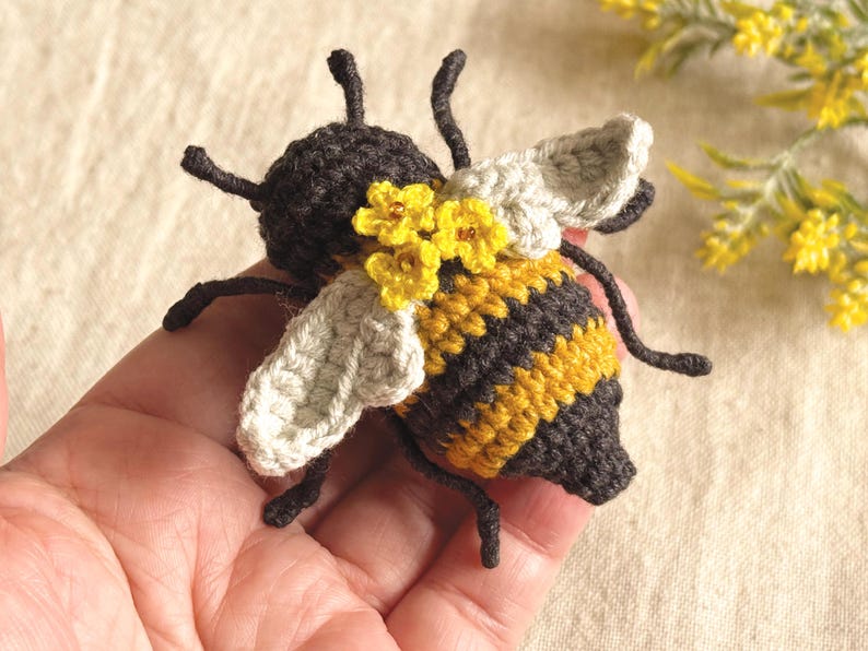 May include: A crocheted bee figurine featuring a dark brown head and tail, with alternating yellow and dark brown stripes on the body. The bee has two light gray crocheted wings and is decorated with three small yellow flowers on its back.