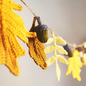 May include: A crochet garland featuring yellow and brown oak leaves and small green and brown acorns strung along a thin twine. The textile ornaments are arranged in a repeating pattern to create a decorative hanging display of autumn foliage.