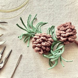 Op de afbeelding: Gehaakte dennenappel- en groenarrangement in bruin- en groentinten. Het handgemaakte ambacht wordt getoond met diverse sieradenmaakgereedschappen, waaronder tangen, scharen en draad, op een neutraal gekleurd stoffen oppervlak.