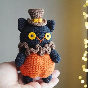 May include: A handmade crochet cat doll with a top hat and orange pumpkin-shaped body. The cat is dark blue with yellow button eyes and a brown ruffled collar. The top hat is brown with a yellow band.