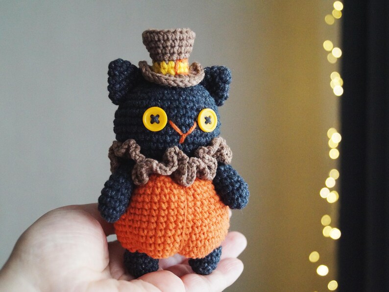 May include: A handmade crochet cat doll with a top hat and orange pumpkin-shaped body. The cat is dark blue with yellow button eyes and a brown ruffled collar. The top hat is brown with a yellow band.