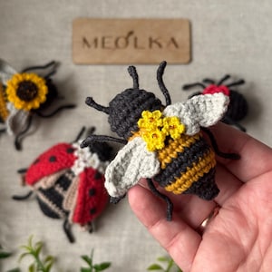 May include: A crocheted bee toy with black and yellow stripes, light grey wings, and small yellow flowers on its back. In the background, there are two other crocheted insects, a ladybird and a sunflower-themed bee, near a wooden sign reading MEOLKA.