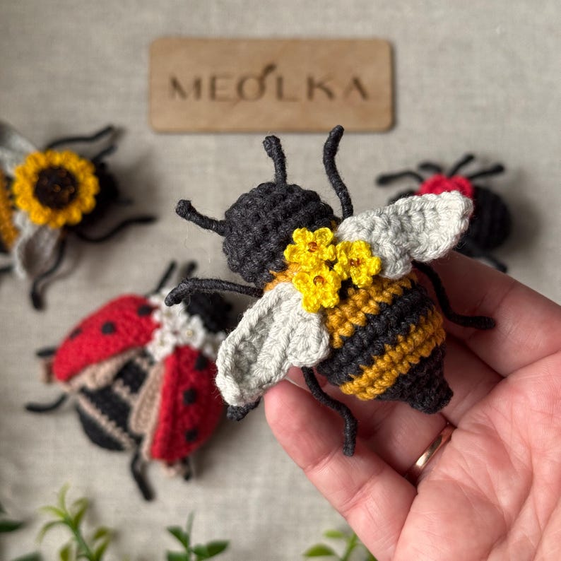 May include: A crocheted bee toy with black and yellow stripes, light grey wings, and small yellow flowers on its back. In the background, there are two other crocheted insects, a ladybird and a sunflower-themed bee, near a wooden sign reading MEOLKA.
