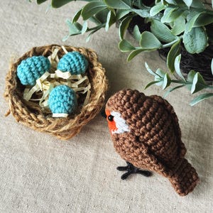 May include: A handmade crochet bird with a brown body, white and orange accents, and black feet. Beside it is a small nest made of twine, containing three turquoise crochet eggs. The scene evokes a springtime theme.
