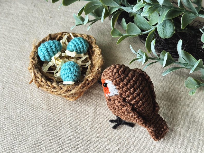 May include: A handmade crochet bird with a brown body, white and orange accents, and black feet. Beside it is a small nest made of twine, containing three turquoise crochet eggs. The scene evokes a springtime theme.