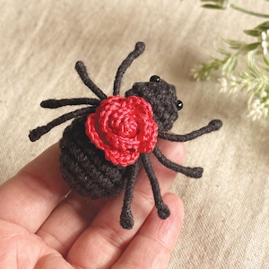 May include: A small, handcrafted crochet spider made from black yarn, featuring eight thin legs and two tiny black bead eyes. A vibrant red crocheted rose is attached to the centre of the spider's back, providing a stark colour contrast.