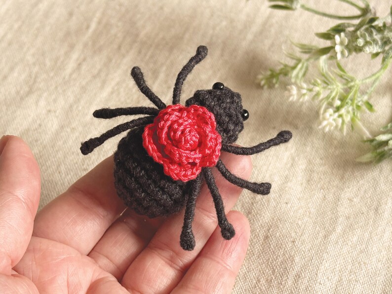 May include: A small, handcrafted crochet spider made from black yarn, featuring eight thin legs and two tiny black bead eyes. A vibrant red crocheted rose is attached to the centre of the spider's back, providing a stark colour contrast.