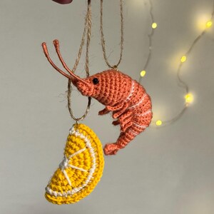 May include: Crocheted ornaments of a shrimp and a lemon slice hanging from twine. The shrimp is orange with white stripes, and the lemon slice is yellow with white details. The background is a soft, neutral color with string lights.