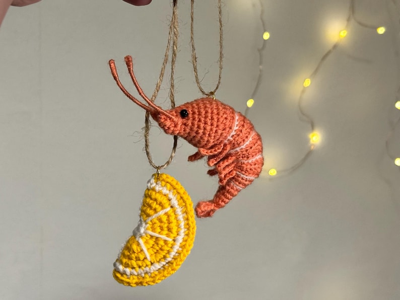 May include: Crocheted ornaments of a shrimp and a lemon slice hanging from twine. The shrimp is orange with white stripes, and the lemon slice is yellow with white details. The background is a soft, neutral color with string lights.