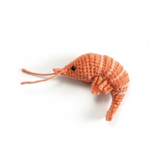 May include: A handmade, orange crochet shrimp with white stripes and black eyes. The shrimp is lying on a white surface. The shrimp's antennae and legs are visible. This item is a unique, handcrafted piece.